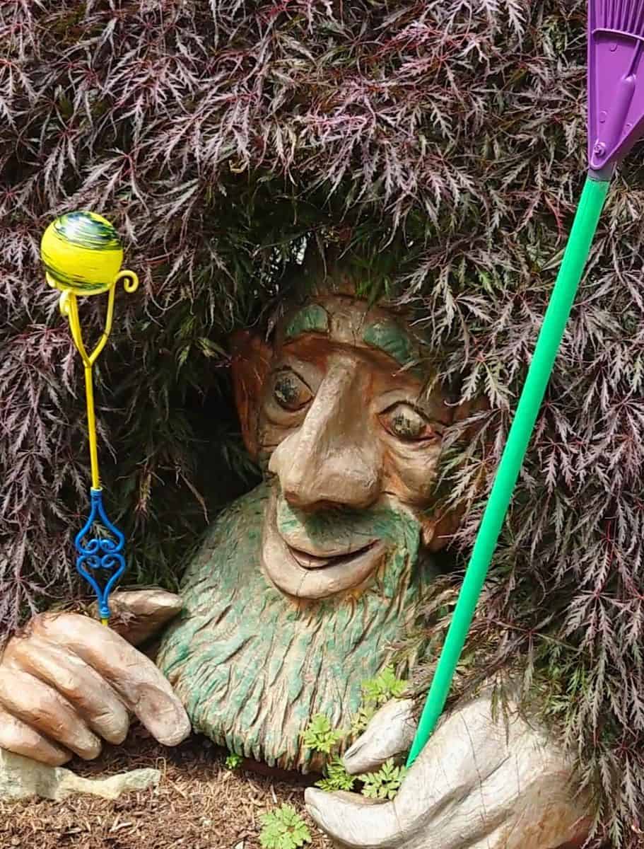 Whimsical troll sculpture with a long nose and green beard peeking out from beneath dense tree branches beside colorful garden decorations. Finding quirky roadside art and hidden sculptures is one of the unexpected things to do in Oak Harbor for visitors exploring the area.