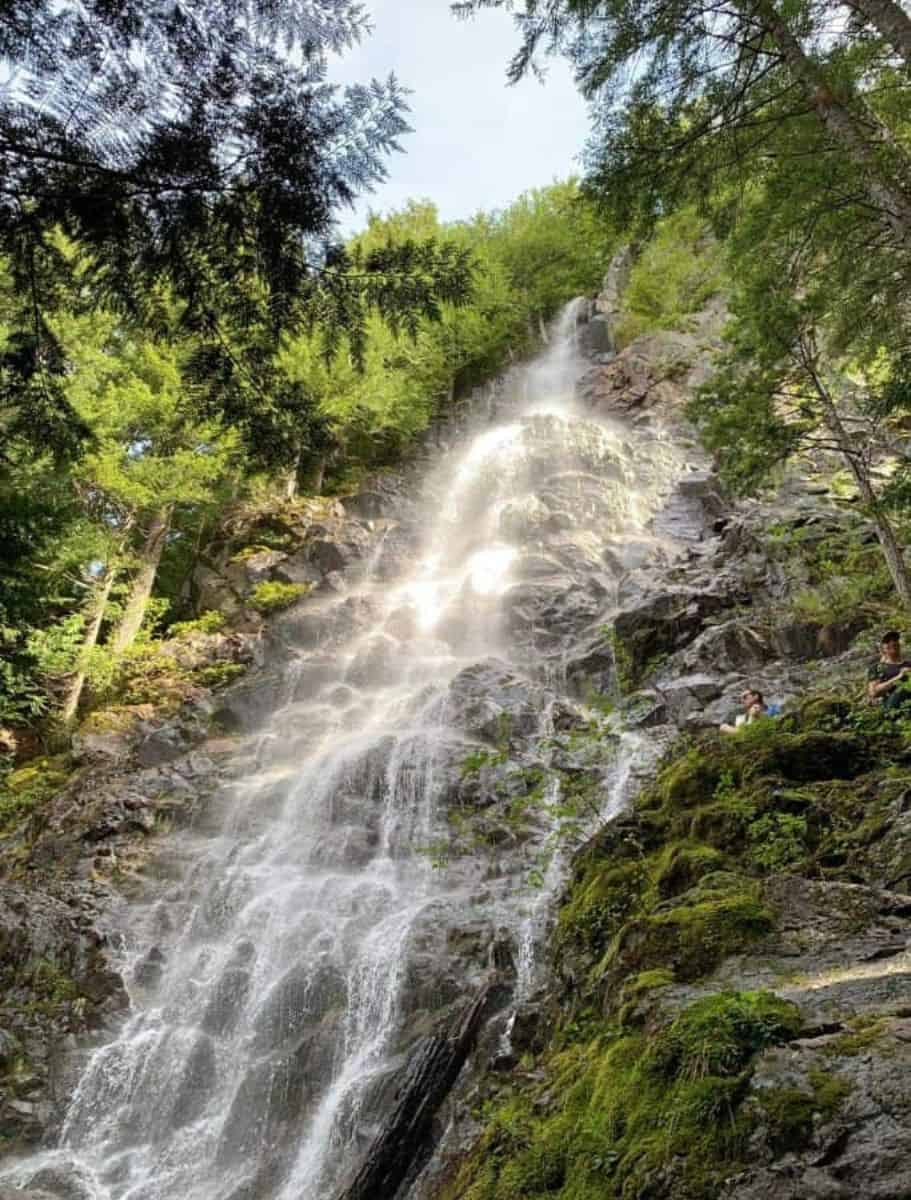 Teneriffe Falls cascading down mossy and rocky cliff in the middle of evergreen forest. people sitting on cliffside halfway up