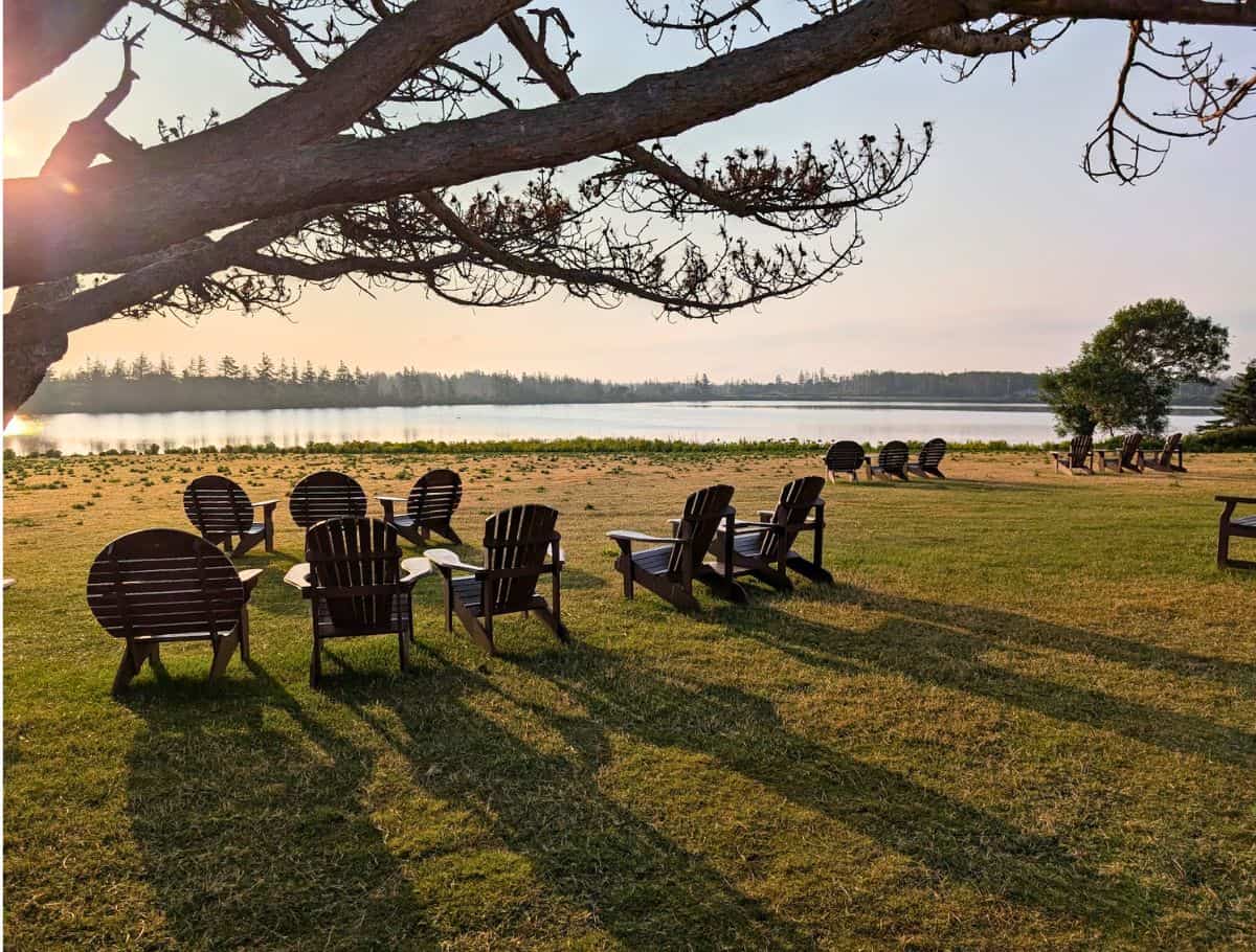 Dalvay by the Sea's Adirondack chairs on the edge of Dalvay Lake with sun peaking through tree in foreground