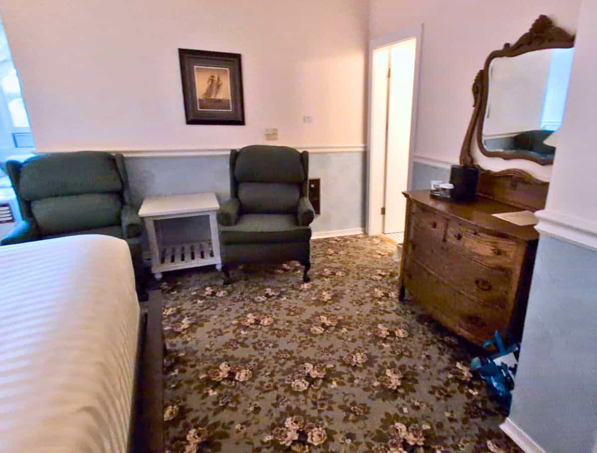 hotel room at the edge of the bed, two wing back chairs, antique dresser with mirror and floral carpet