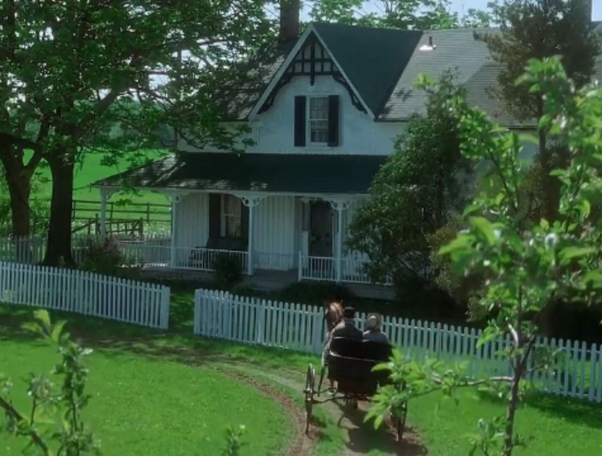 Scene of Anne first arriving by carriage at Green Gables house surrounded by white picket fence and vibrant greenery