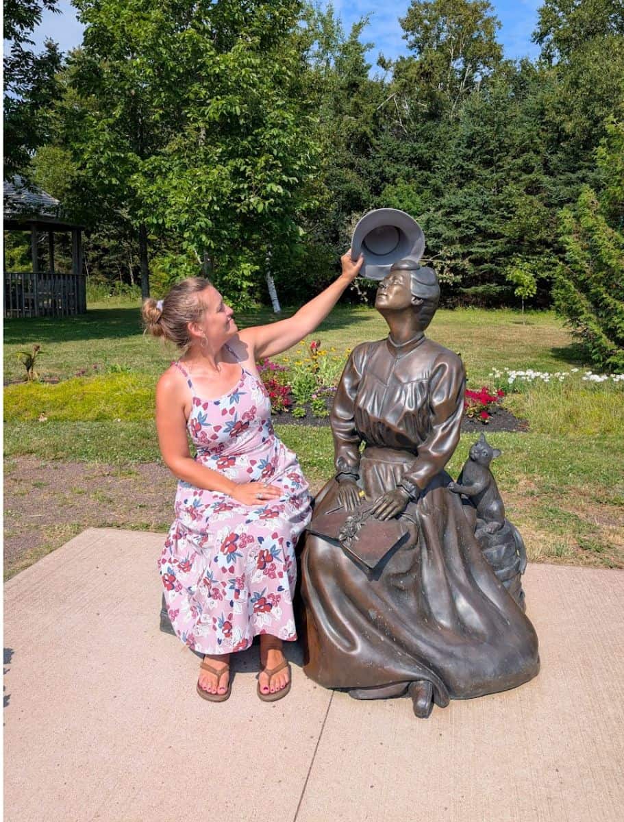 Me giving L.M. Montgomery statue my hat. she is sitting on bench, eyes closed, head tilted to sky