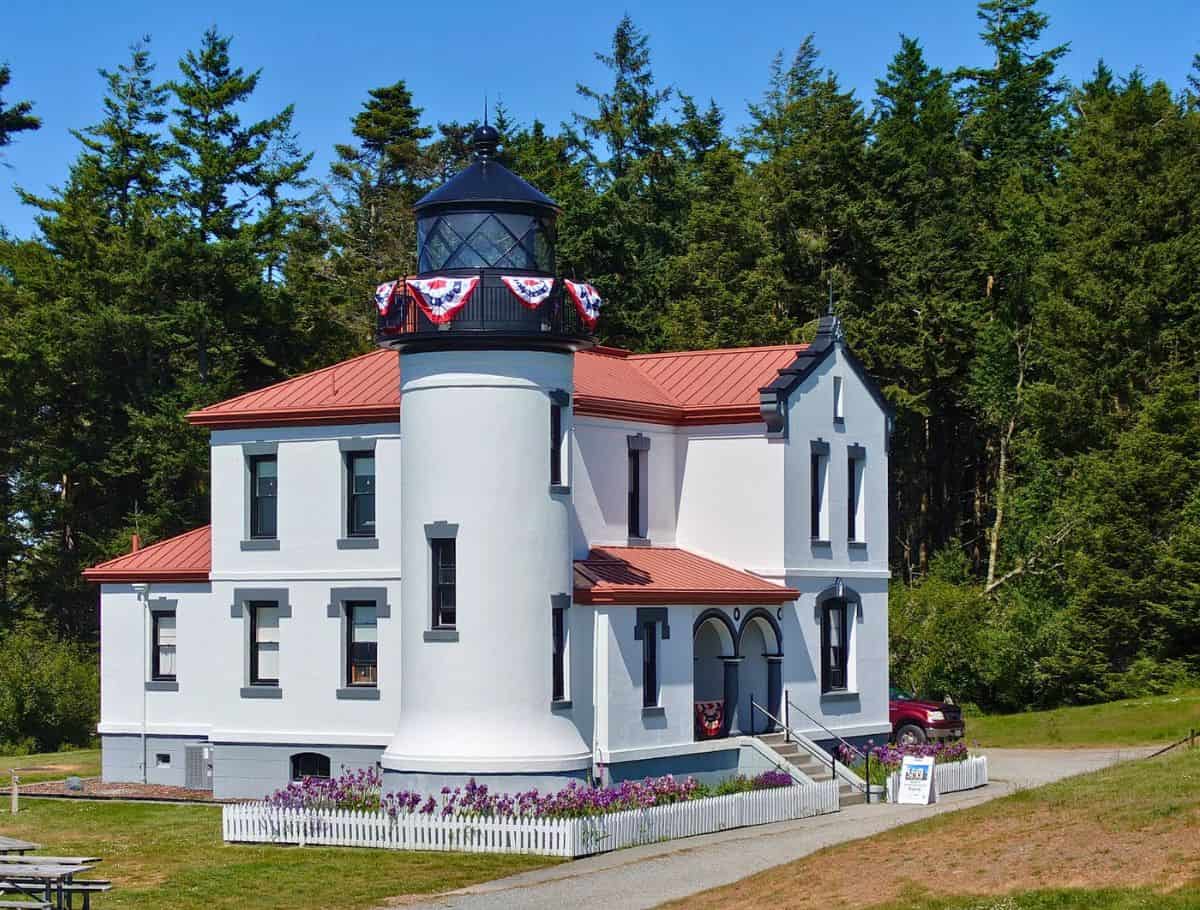 White lighthouse with a red roof and black lantern sits among tall evergreen trees with patriotic bunting decorating the balcony. This historic landmark adds a classic sightseeing stop during a weekend on Whidbey Island.