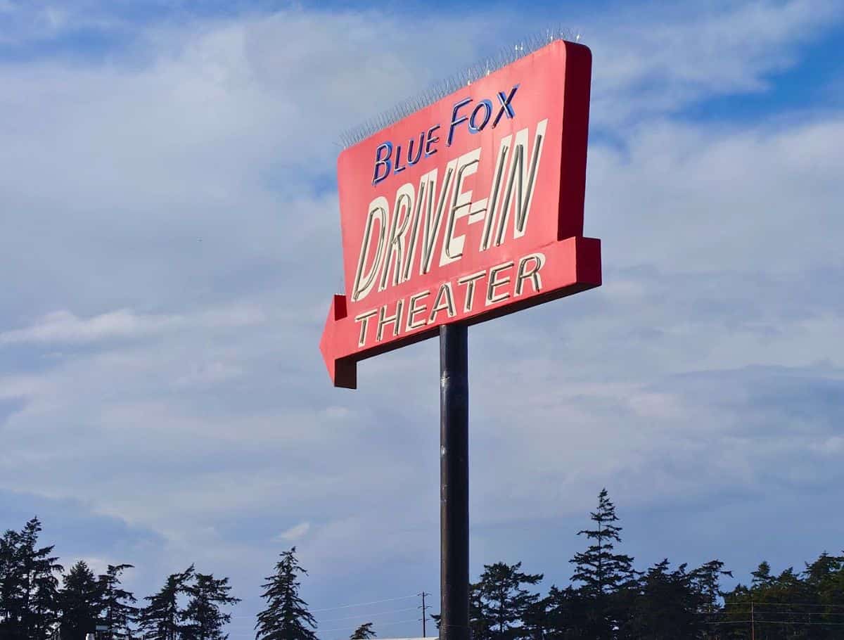 Retro roadside sign reading "Blue Fox Drive In Theater" in bold red and white lettering against a partly cloudy sky. Catching a movie at this nostalgic drive in is one of the fun and unique things to do Whidbey Island visitors can enjoy.