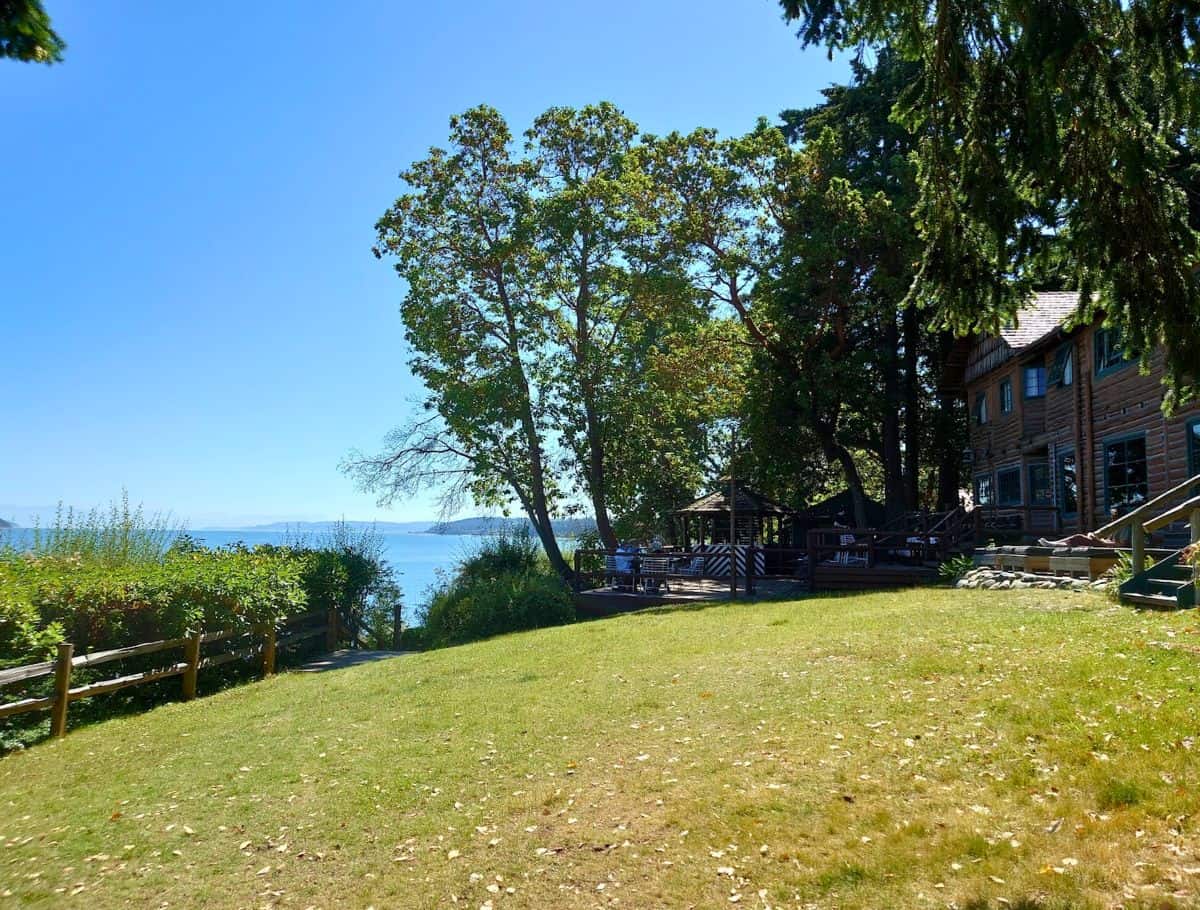 Sunny waterfront lawn with a rustic wooden building and shaded outdoor seating overlooking calm blue water and distant shoreline. Relaxing at scenic waterfront spots like this is one of the peaceful things to do Whidbey Island visitors can enjoy.