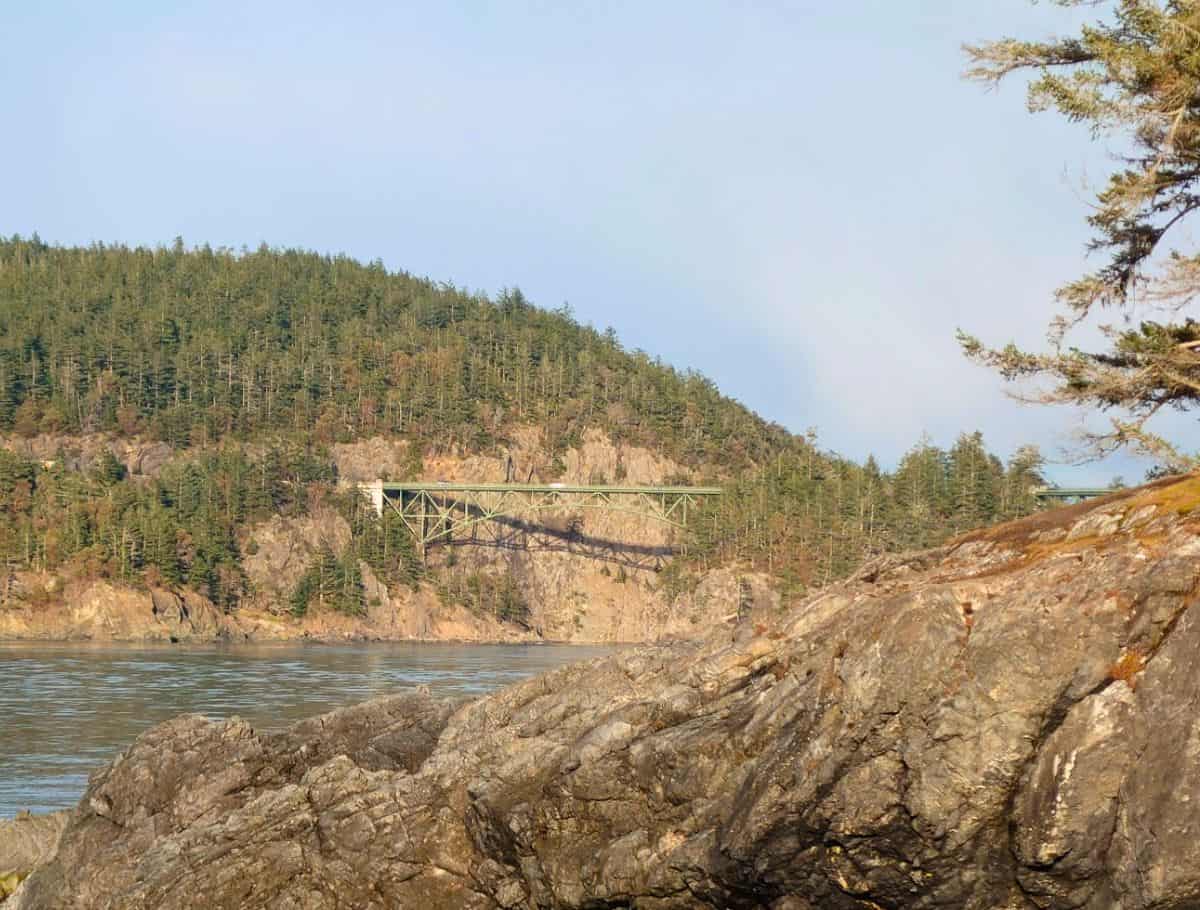 Rocky shoreline in the foreground with calm water leading to a forested cliff and a narrow green bridge spanning the gap. This scenic coastal view highlights one of the peaceful outdoor things to do Whidbey Island visitors can enjoy while exploring the shoreline.
