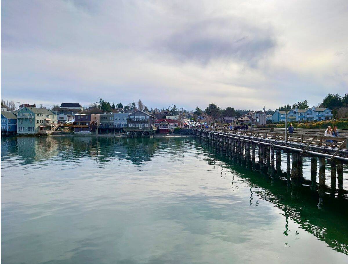 Wooden pier lined with people stretches over calm water toward colorful waterfront buildings in a small coastal town under a cloudy sky. Strolling along charming seaside towns like this is one of the relaxing things to do Whidbey Island visitors can enjoy.
