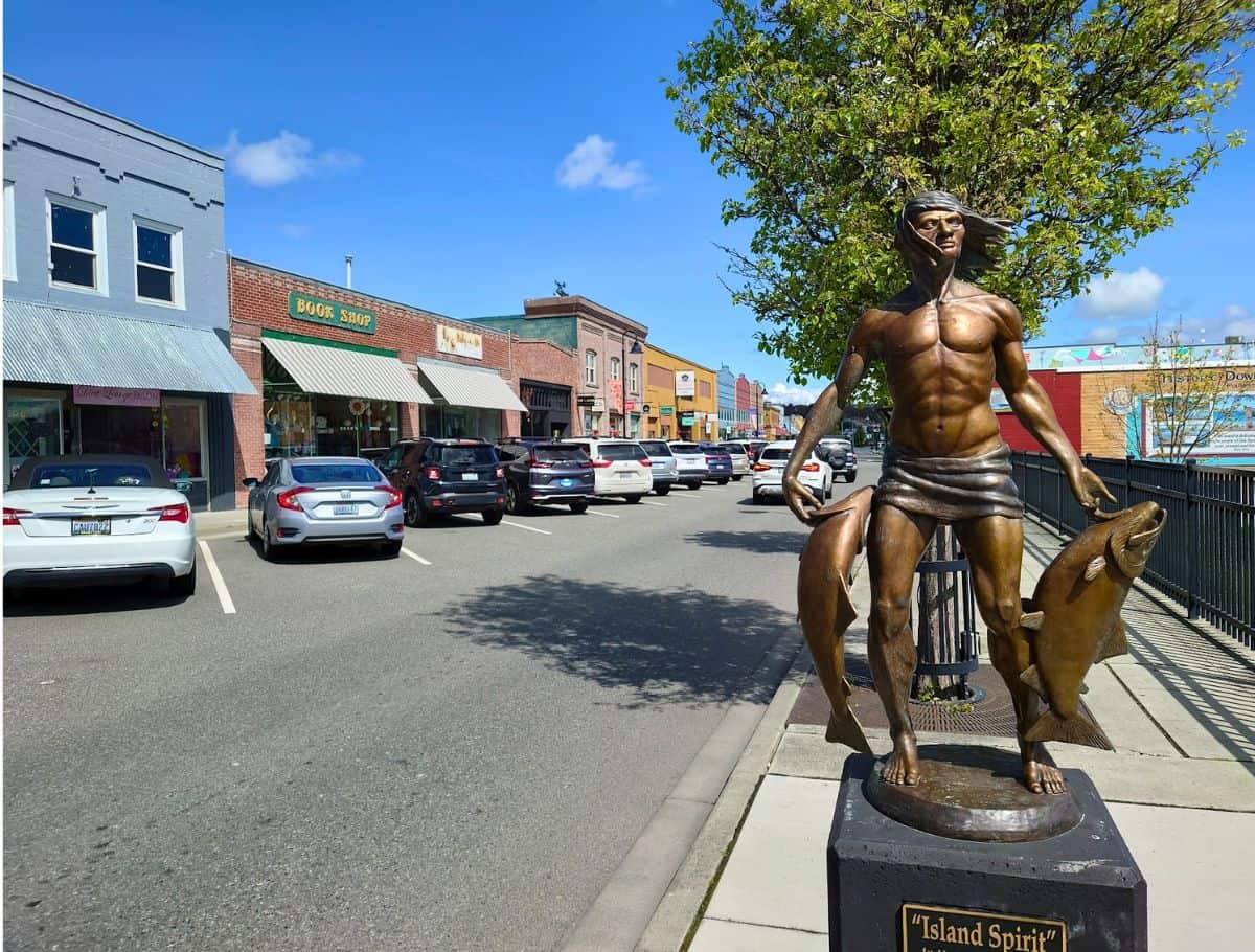 Bronze statue of a muscular figure holding fish stands along a small town street lined with colorful storefronts and parked cars under a bright blue sky. The statue base reads "Island Spirit" and marks a charming downtown stop to explore during a weekend on Whidbey Island.