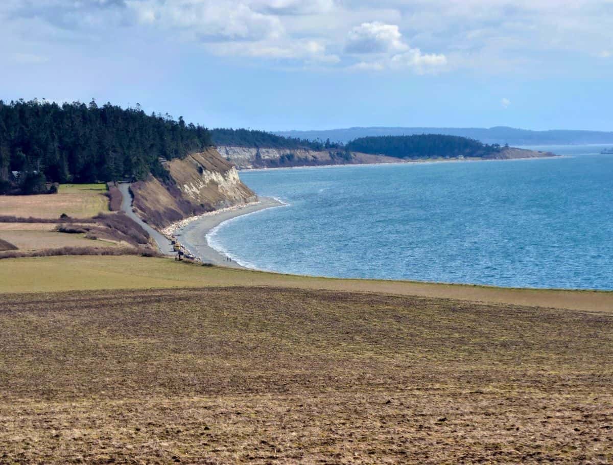 Sweeping coastal view of rolling farmland leading to dramatic cliffs and a curved shoreline with calm blue water under a partly cloudy sky. Taking in scenic viewpoints like this is one of the most breathtaking things to do Whidbey Island visitors can experience.