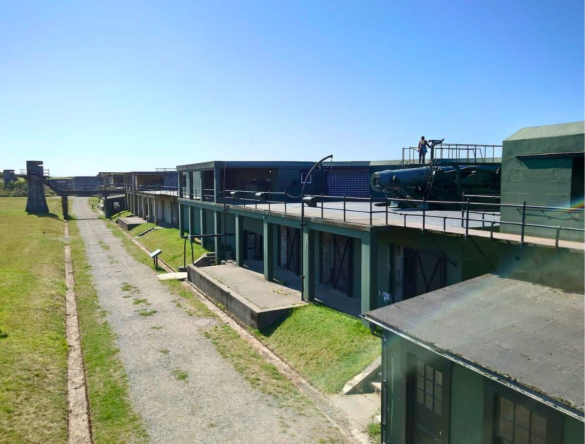 Historic coastal fort with long green buildings, mounted artillery, and a gravel path stretching alongside open grassy grounds under a clear blue sky. Exploring this preserved military site is one of the more unique and educational things to do Whidbey Island visitors can experience.