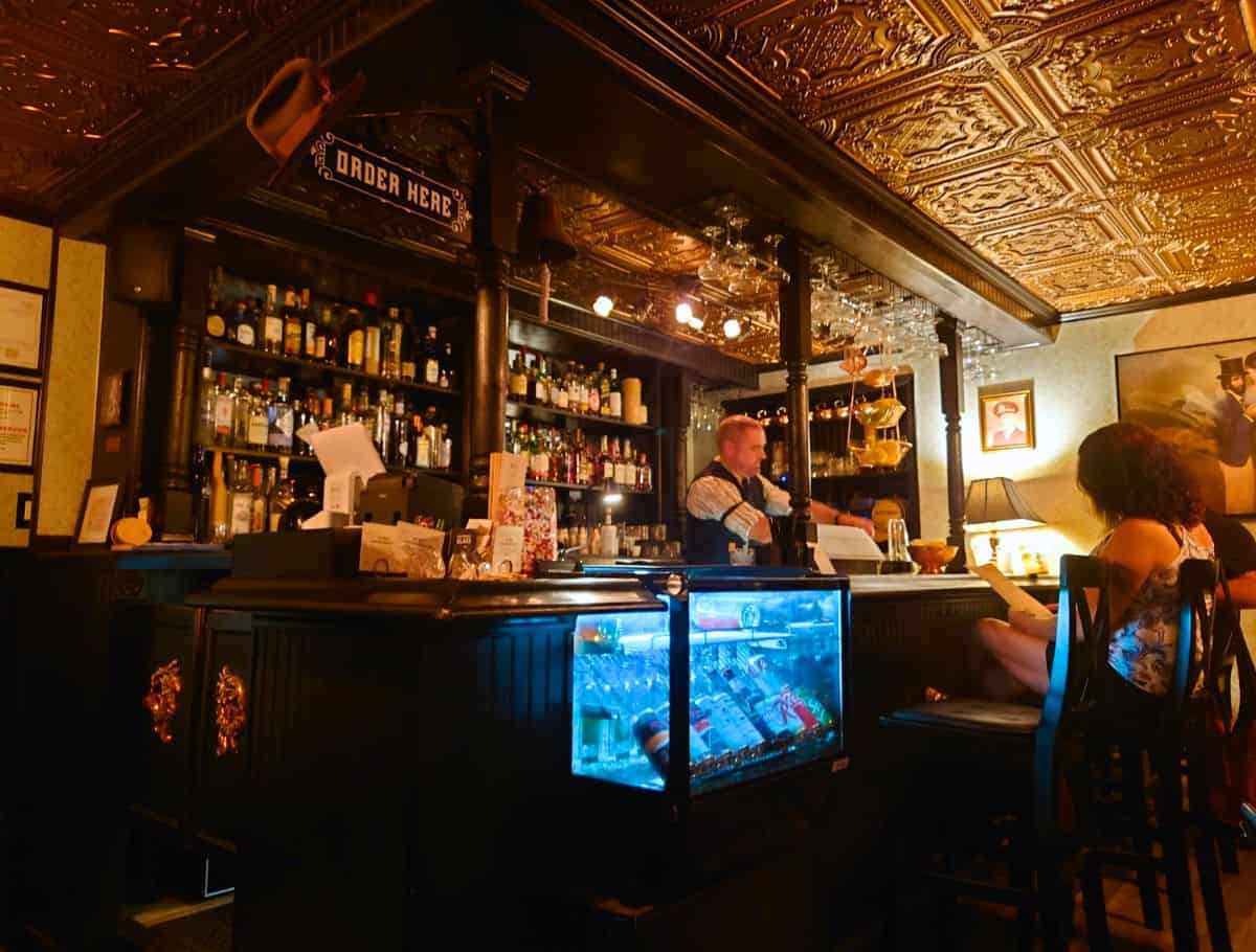 Cozy vintage style bar in Langley with a bartender serving drinks behind a dark wood counter beneath a decorative pressed tin ceiling, offering a lively nightlife experience among things to do Langley. Shelves of liquor bottles, hanging glassware, and warm lighting create an intimate atmosphere while a guest sits at the bar reading a menu.