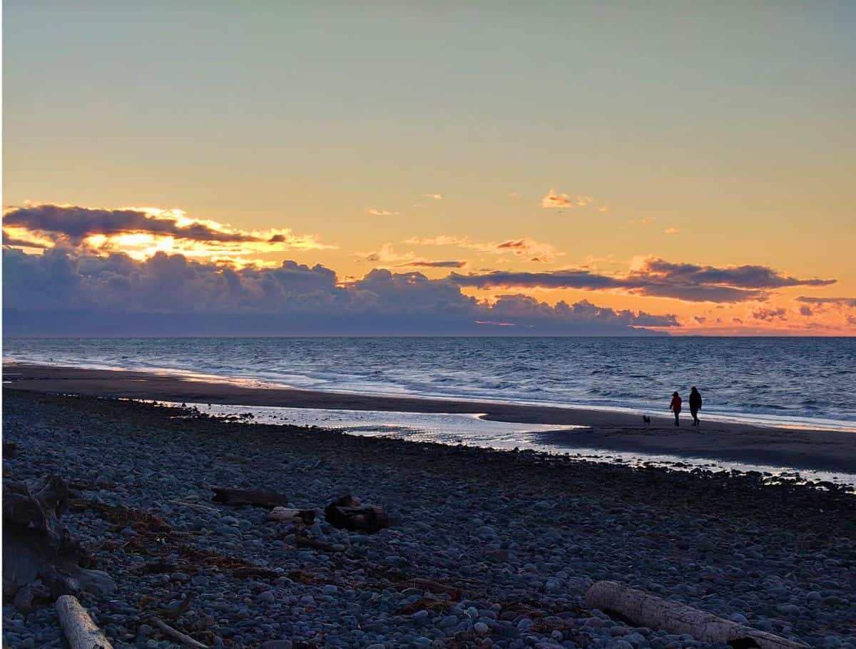 Two people walk along a rocky beach at sunset with a small dog as waves roll in under a glowing orange and blue sky. This quiet shoreline moment captures the relaxed pace of a weekend on Whidbey Island.