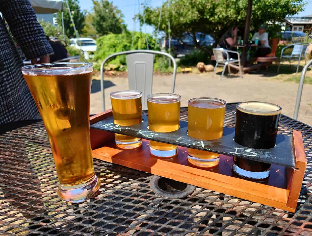 Flight of craft beers in small glasses and a full pint sit on a metal patio table at an outdoor brewery with people relaxing in the background. This casual tasting experience highlights a fun way to unwind during a weekend on Whidbey Island.