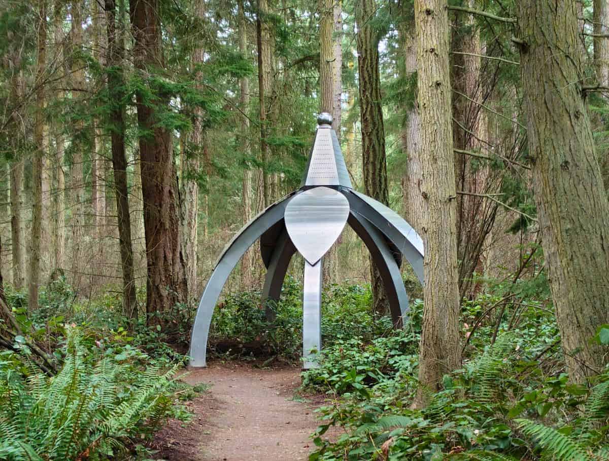 Metal sculpture shaped like an open dome stands along a forest trail surrounded by tall trees and green ferns. This artistic installation adds a unique and peaceful stop to explore during a weekend on Whidbey Island.