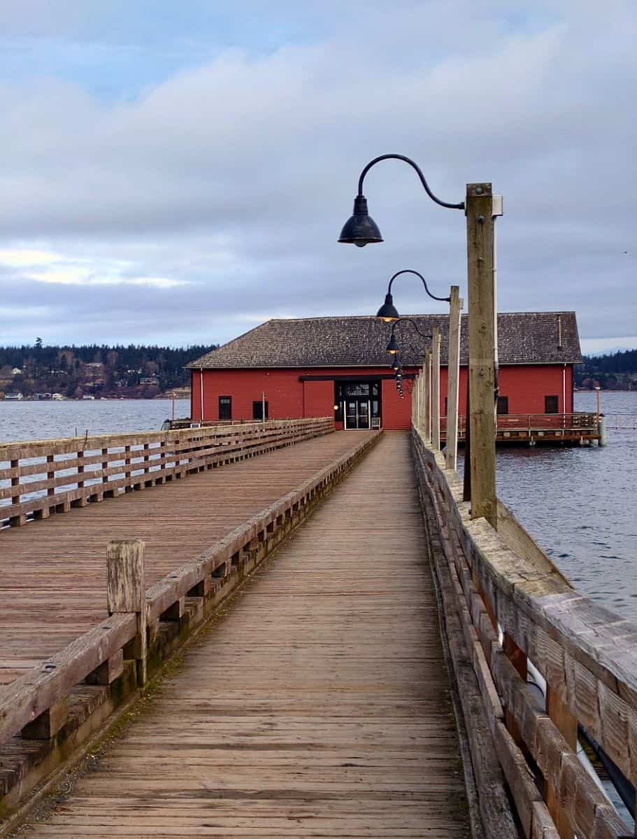 Wooden pier lined with curved lampposts leads to a red waterfront building over calm water beneath a cloudy sky. This inviting walkway showcases a scenic spot to visit during a weekend on Whidbey Island.