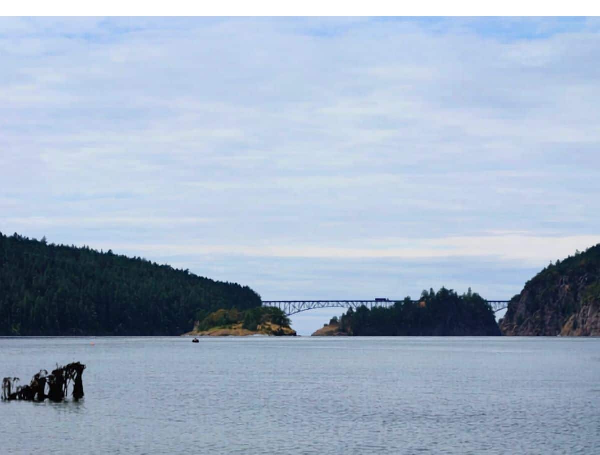 Calm coastal water framed by forested shorelines with a long bridge stretching between two land masses under a cloudy sky. This peaceful waterfront view highlights a scenic stop to enjoy during a weekend on Whidbey Island.