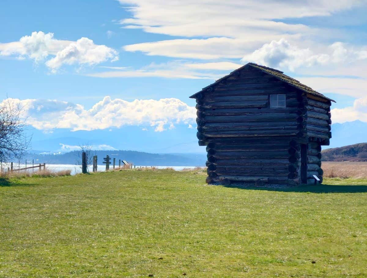 Small rustic log cabin sits alone in a grassy field overlooking water and distant mountains beneath a bright blue sky with scattered clouds. This quiet historic scene reflects the slower pace and scenic charm of a weekend on Whidbey Island.