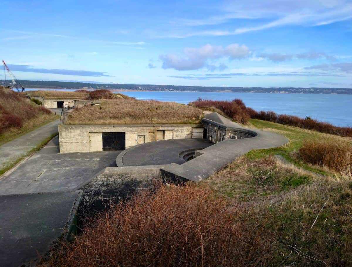 Concrete coastal fort structure with curved walls sits on a grassy bluff overlooking calm blue water and distant shoreline under a partly cloudy sky. This historic site offers sweeping views and a glimpse into the past during a weekend on Whidbey Island.