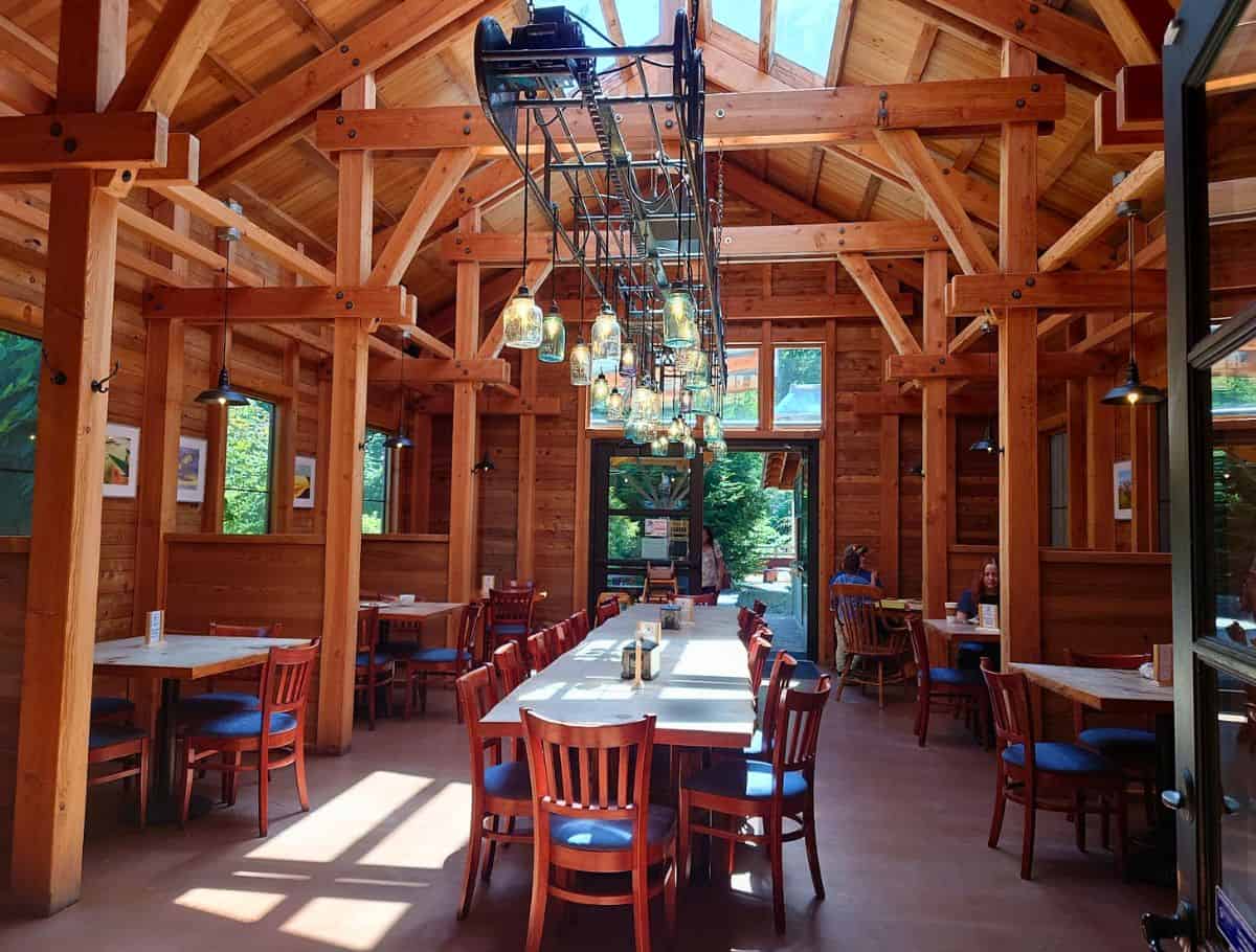 Warm wood interior of a cozy restaurant in Langley with long communal tables, wooden beams, and hanging jar lights creating a welcoming dining space among things to do Langley. Sunlight pours in through large windows while a few guests sit and enjoy a relaxed meal.