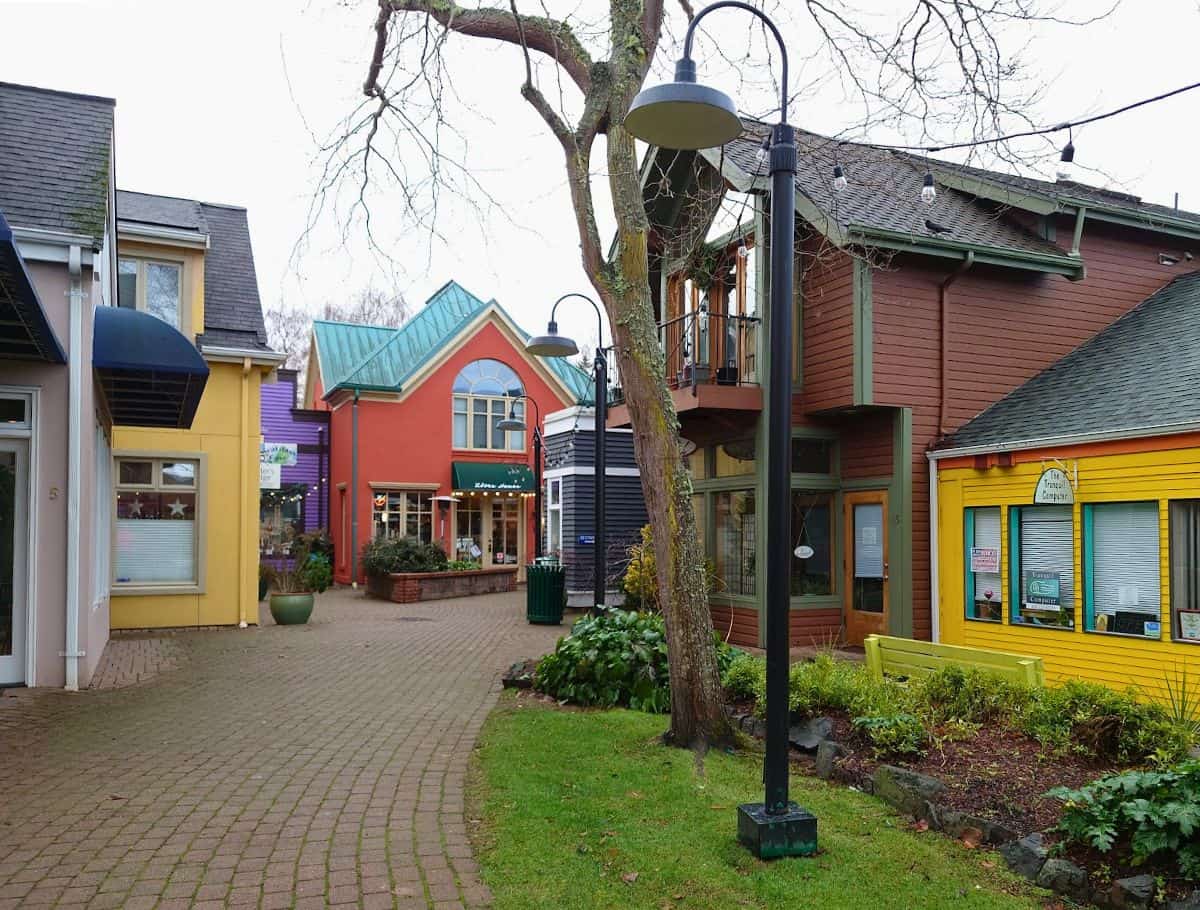 Colorful storefronts and small shops line a quiet pedestrian walkway in Langley, with bright yellow, red, and purple buildings creating a charming village feel for visitors exploring things to do Langley. A tree and street lamps frame the path, adding to the relaxed and walkable atmosphere.