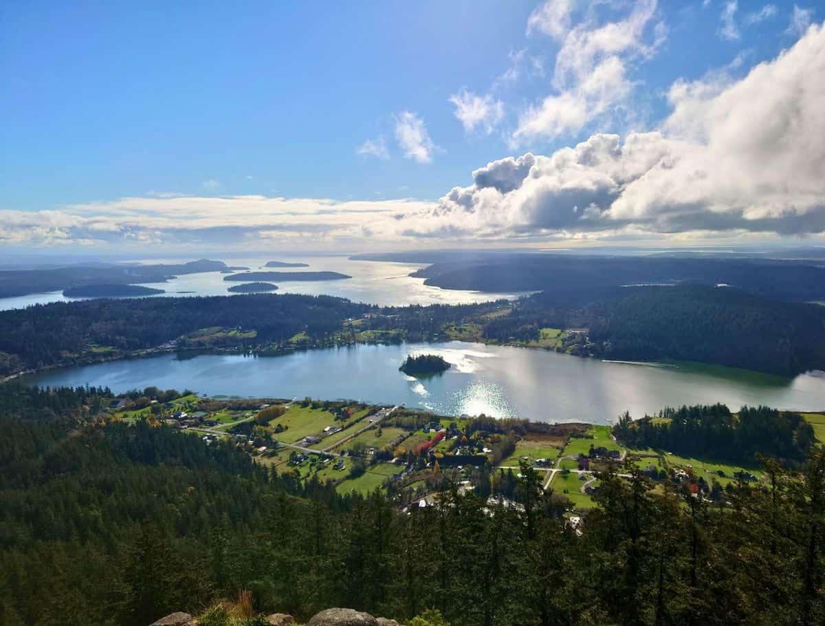 Sweeping view from above of a calm lake surrounded by forest and scattered homes, with sunlight reflecting off the water and distant islands under a dramatic sky. This scenic overlook captures the peaceful landscape you might experience during a weekend on Whidbey Island.
