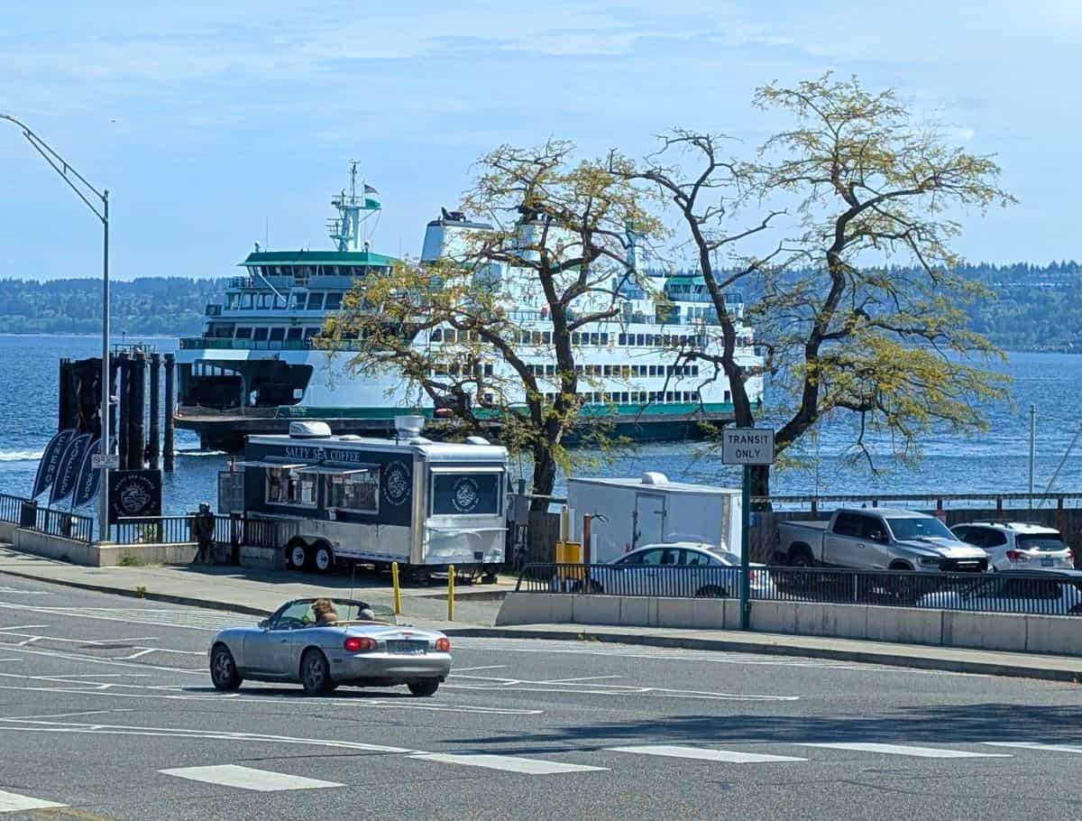 Ferry boat docks along the waterfront behind a small coffee stand labeled "SALTY SEA COFFEE" with cars passing by in the foreground. This arrival scene captures the start of a weekend on Whidbey Island with easy access by ferry and local stops nearby.