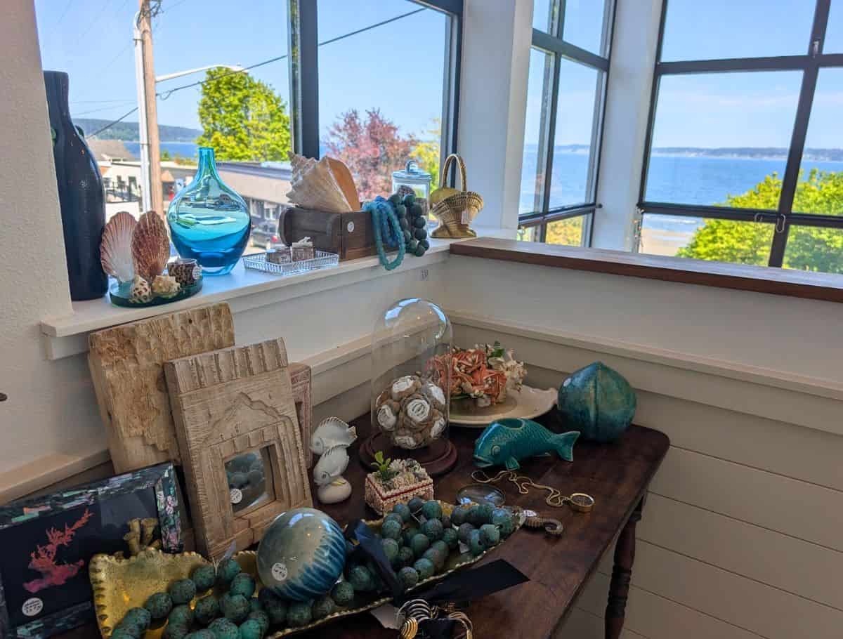 Coastal themed shop display with seashell decor, glass vases, and turquoise accents arranged on a wooden table by a bright window overlooking the ocean, highlighting scenic and relaxing things to do Langley. Sunlight streams in, revealing a peaceful waterfront view beyond the curated home goods.
