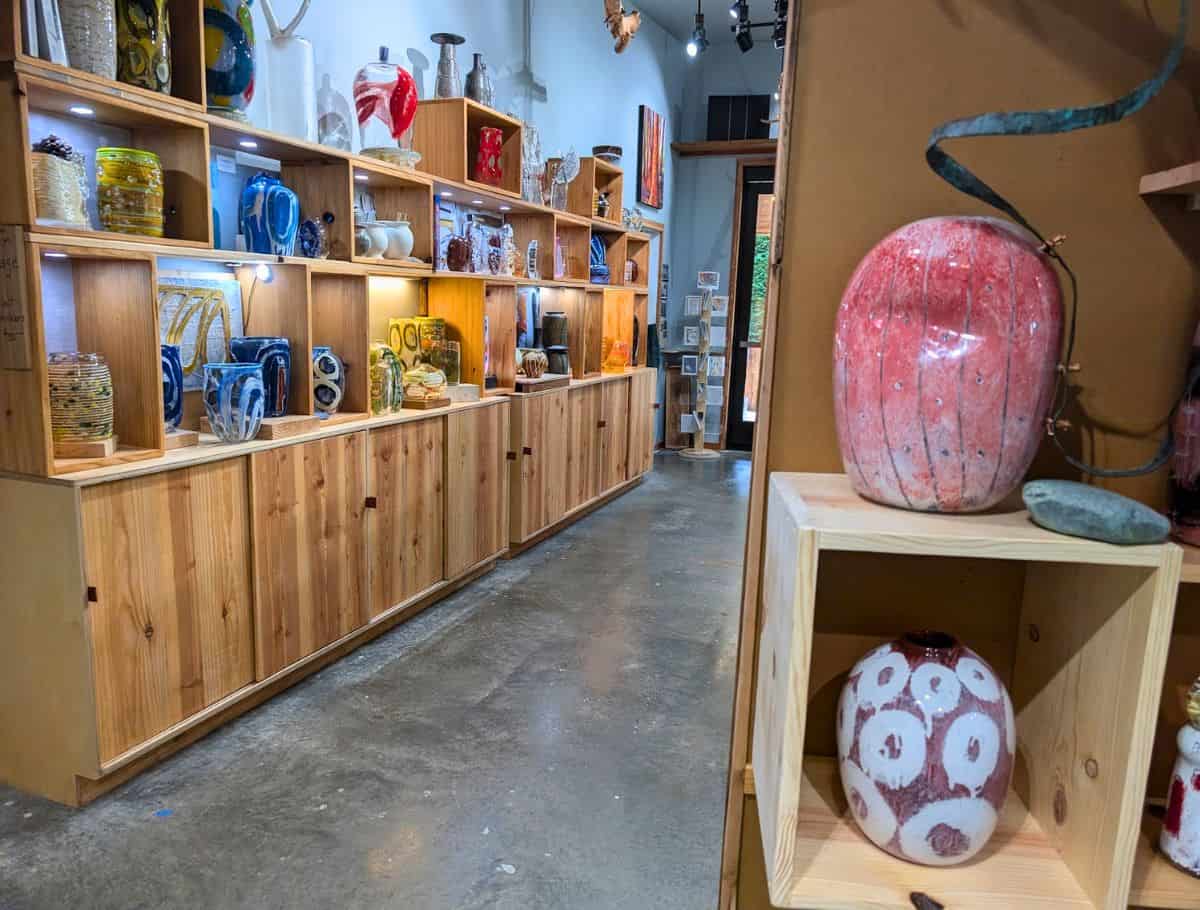 Bright art gallery in Langley displaying colorful glass vases and handmade pieces arranged on wooden shelves, highlighting a creative stop among things to do Langley. The space features warm lighting and a modern rustic interior that invites visitors to browse local artwork.
