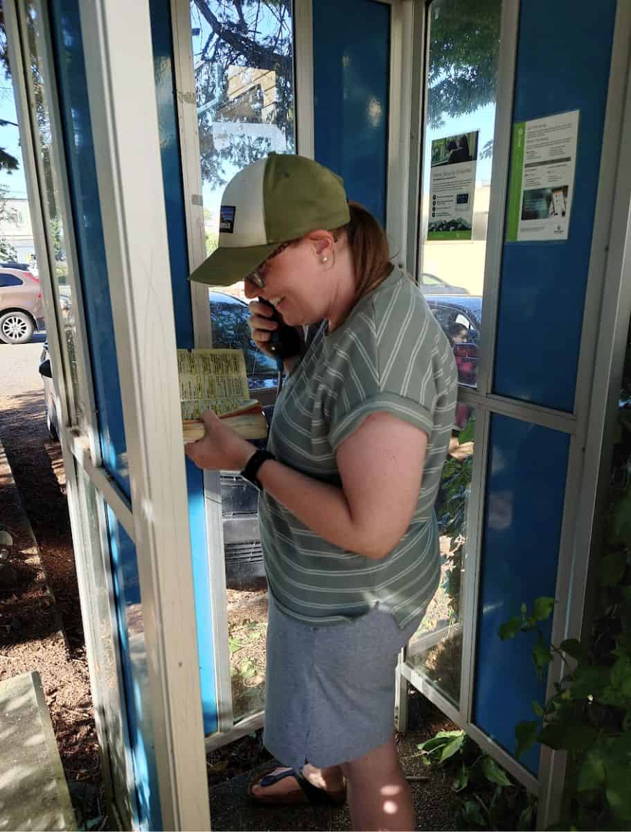 Woman smiling while using a public phone booth and flipping through a directory, capturing a quirky retro activity among unique things to do Langley. She wears a green cap and striped shirt, standing inside a blue booth with sunlight filtering through the windows.
