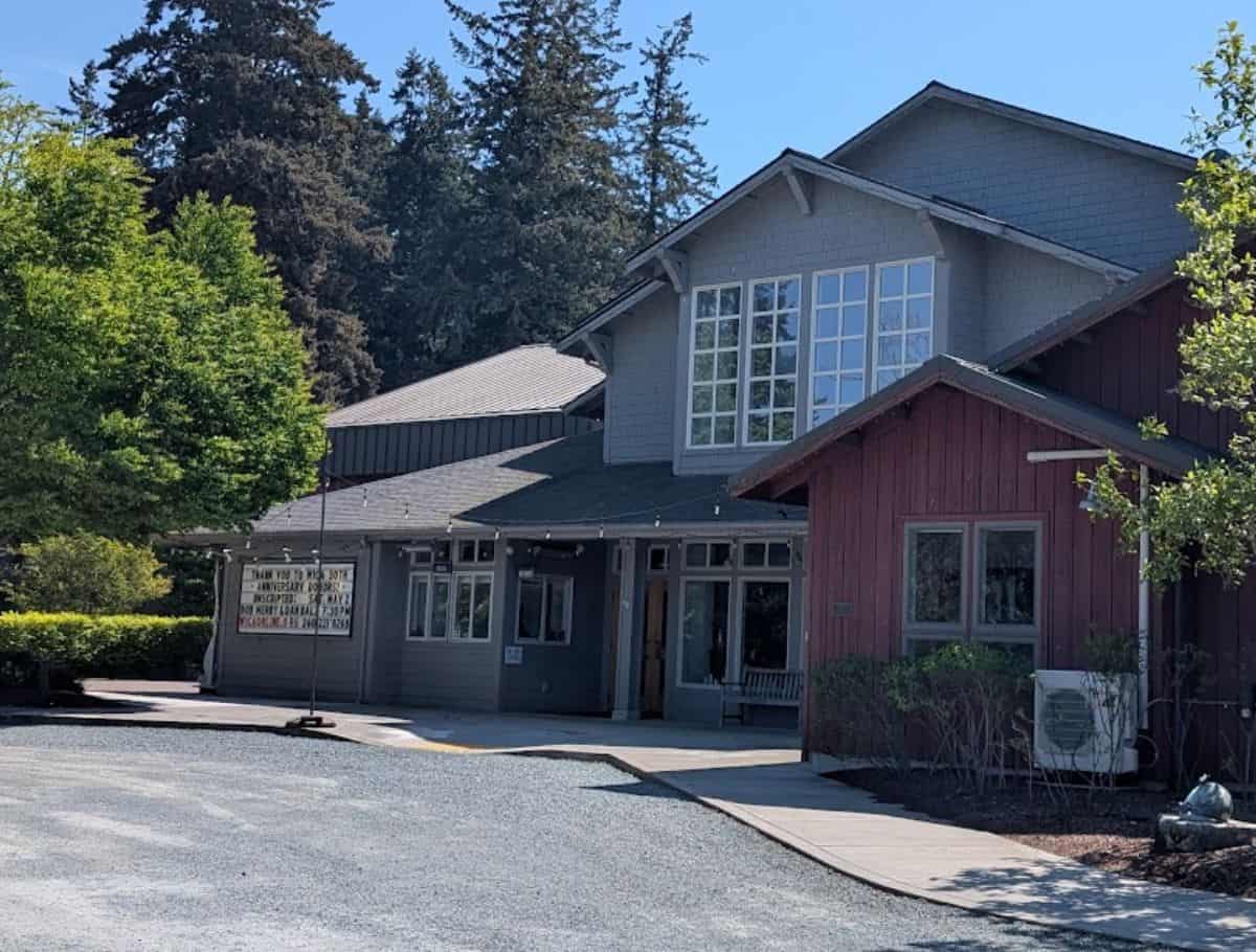 Charming community building in Langley surrounded by trees with a welcoming entrance and event sign out front, representing a local venue to check out when exploring things to do Langley. The mix of grey and red siding and large windows gives it a cozy small town feel.
