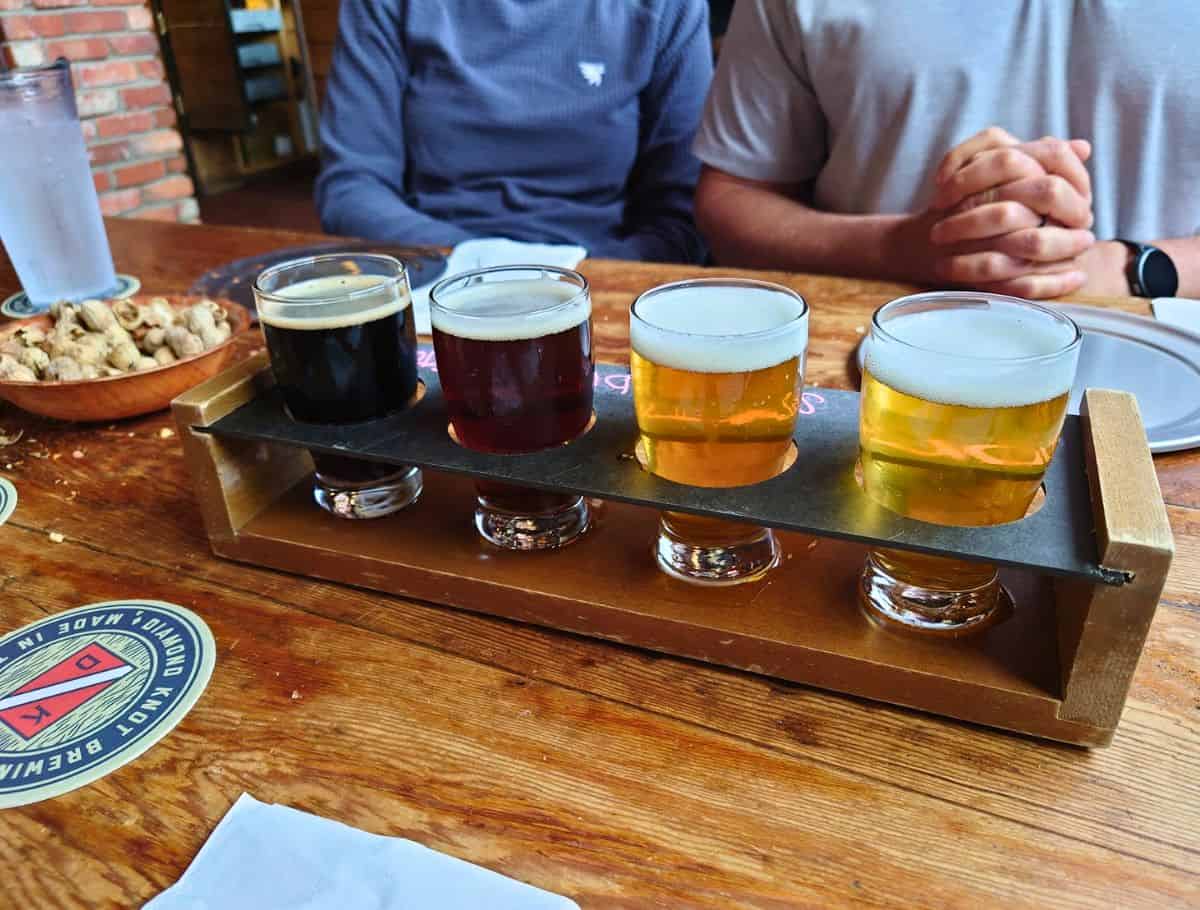 Beer flight with four small glasses ranging from dark stout to light lager served on a wooden paddle at a rustic table, a popular tasting experience and one of the things to do Langley. Two people sit behind the drinks with snacks and water on the table, creating a relaxed brewery setting.