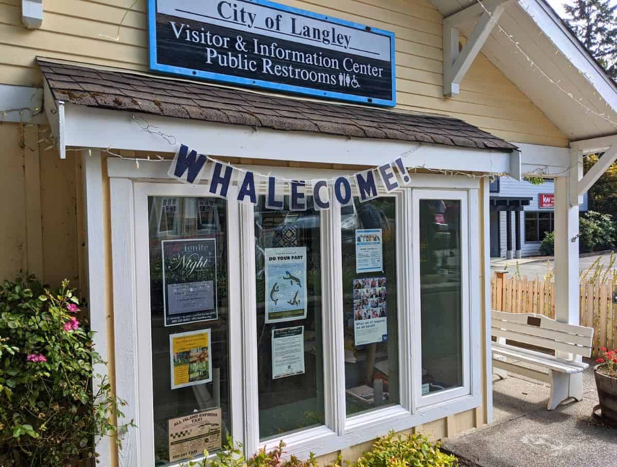 Small yellow visitor center building with a sign reading "City of Langley Visitor and Information Center Public Restrooms" and a banner that says "WHALECOME!" hanging above the windows, showcasing a helpful stop when exploring things to do Langley. Posters and local notices fill the windows, adding context for visitors looking for activities and information.