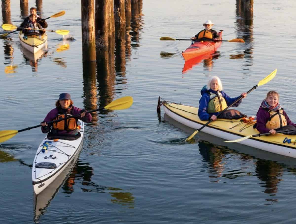 Group of people kayaking through calm water near wooden pilings in Langley, showcasing an adventurous outdoor activity among things to do Langley. Brightly colored kayaks and paddles reflect on the water as paddlers explore the shoreline together.